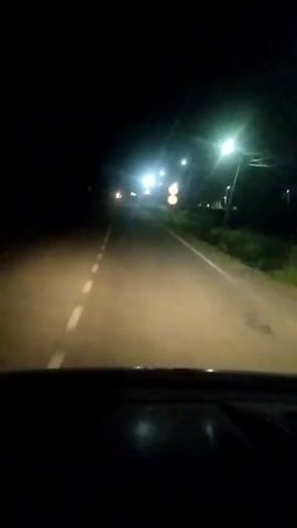 A first-person perspective video shows a car driving on a road at night. Streetlights illuminate the road and the surrounding dark environment. The car's headlights cast a beam of light on the asphalt, which has a white line marking the center. In the distance, several other headlights are visible, suggesting other vehicles on the road. A speed limit sign indicating 50 is visible on the right side of the road. The dashboard of the car is visible in the foreground, partially obscured by darkness. The overall atmosphere is dark and quiet, with the focus on the journey ahead.