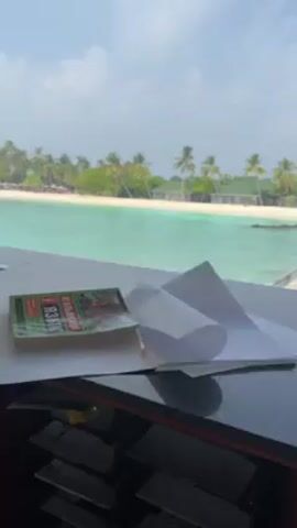 A static, eye-level shot reveals a tropical beach setting through an open window, contrasted with a cluttered foreground. The beach features turquoise water, a white sandy beach, palm trees, and small structures, suggesting a resort. In the immediate foreground, a counter or tabletop is visible, holding a packet of cigarettes, papers, and other unidentified items. The papers are crumpled and partially obscured, giving the impression of a workspace in use. The background light washes out the foreground details, leaving the exact nature of the desk items somewhat vague. The overall impression is of a work or resting area overlooking a beautiful tropical landscape.