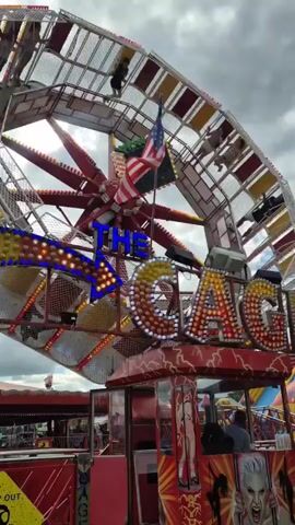 The video shows a rotating amusement park ride called "The Cage" in operation. The ride consists of a central rotating arm with a large circular cage attached to the end.  People are riding inside the cage as it spins and rotates. The ride is decorated with lights, flags, and signage. The sky is cloudy, and other carnival attractions are visible in the background. The camera angle is looking up at the ride from a ground-level perspective.