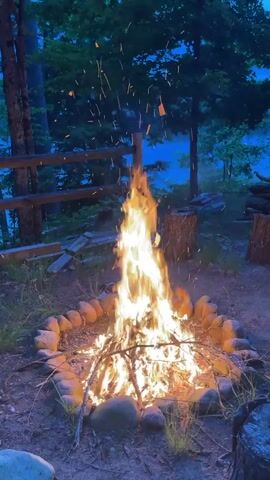The video shows a campfire burning in a stone-lined pit. The fire is tall and bright, with sparks rising into the air. Behind the fire, a wooden fence and trees are visible, with a body of water in the distance. The scene is dark, suggesting it is evening or night. There are no people or animals visible in the video. The ground around the fire pit is covered in dirt and dry leaves.