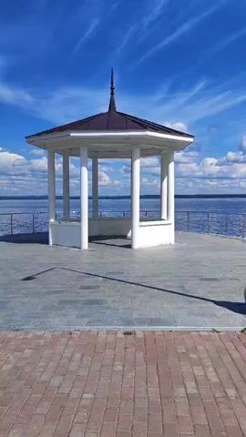 A white gazebo with a dark roof and a pointed finial sits on a paved area near a body of water. The gazebo has eight white columns and a low white wall around its base. The ground in front of the gazebo is paved with large gray tiles, and a path of reddish-brown bricks runs along the foreground. Beyond the gazebo, a railing separates the paved area from the water, which extends to the horizon. The sky above is a bright blue with scattered white clouds. The sun casts a long shadow of the gazebo onto the paved ground. There are no people visible in the video.