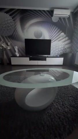 This is a still image of a modern living room. The main subjects are the furniture and the room's decor. In the foreground, a white, oval-shaped coffee table with a thick, clear glass top rests on a dark gray, textured rug. Behind the coffee table, a long, white entertainment unit with dark accents supports a large, flat-screen television. The wall behind the entertainment unit is decorated with a striking mural featuring a black and white, abstract, swirling pattern that resembles a vortex or a spiraling galaxy. There are also textured, geometric decorative elements on the wall, resembling spheres or starbursts. To the left of the television, a tall, light-colored vase holds artificial white flowers. Next to the vase, a white abstract sculpture stands on the entertainment unit. To the right of the television, a wall-mounted air conditioning unit is visible near the ceiling. The overall aesthetic is sleek, contemporary, and monochromatic, with a strong emphasis on geometric shapes and abstract art. There are no people visible in the image. The lighting suggests it is daytime, likely indoors. No weather is discernible. The image appears to be taken in a residential setting, likely a living room in Izmail, Ukraine. There is no discernible text in the image.