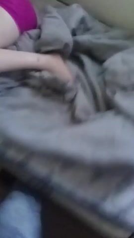 A blurry, close-up video shows a person lying face down on a bed with a gray quilted bedspread. They are wearing bright pink shorts and a black top. A hand enters the frame and pats the person's bottom before the video goes dark.