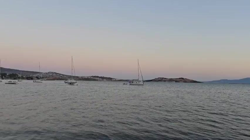 The video shows a wide shot of a body of water with several sailboats anchored. The sky is a pale pink and purple, indicating either sunrise or sunset. In the distance, there is a hilly coastline with some buildings visible. The water is calm with gentle ripples. There are no people clearly visible in the frame, and no actions or interactions are taking place. The overall scene is serene and tranquil.