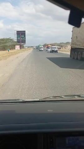 The view from inside a car driving on a highway. The road is straight with light traffic including cars, vans, and large trucks. There is a billboard on the left side of the road with text on it. The sky is partly cloudy.