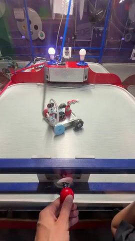A person's hand is holding a red ball near a small robot on a silver surface. The robot has wheels and some electronic components. The scene appears to be some kind of game or exhibit. The robot moves and appears to interact with the ball, pushing it. The background shows a colorful environment with lights and displays.