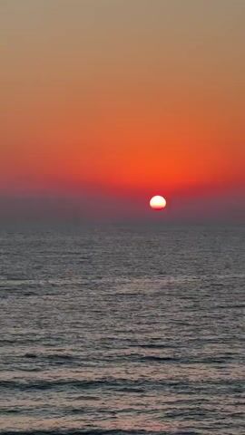 A vertical video captures a sunset over the ocean. The sun is a small, bright orb just above the horizon, casting a reddish-orange glow across the sky and reflecting on the water. Gentle waves create a textured surface on the sea. The sky transitions from a deep orange near the horizon to a lighter, gradient hue towards the top of the frame.