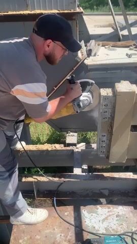 A man wearing a grey t-shirt, grey pants with a white stripe, a black baseball cap, and white sneakers is operating a large yellow angle grinder. He is seen from the side, facing a concrete block that is being cut. The grinder is creating a dust cloud as it cuts into the block, which is secured in place on a metal structure. In the background, there is more industrial-looking equipment and some greenery. The man appears focused on his task, with no overt emotional expression. The scene suggests an outdoor, possibly industrial or construction setting. A black power cord runs from the grinder to an unseen power source. A green power tool, a Makita, is visible at the bottom right of the frame. There is no visible text in the media. The lighting suggests it is daytime, likely sunny, given the bright highlights and shadows.