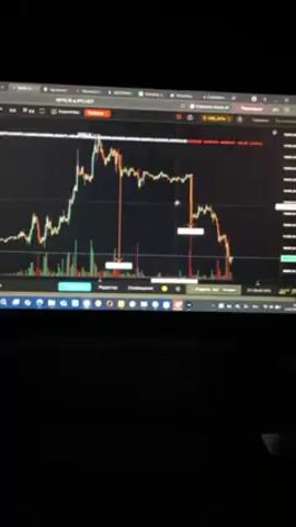 The video shows a laptop screen displaying a financial graph, presumably related to cryptocurrency or stock trading. The graph has a black background with red and green bars representing market fluctuations. There are two vertical red arrows indicating specific points on the graph, possibly highlighting price drops or significant events. The laptop keyboard is visible in the foreground, partially obscured by darkness. The overall setting is dimly lit, drawing focus to the screen's display. The focus is on analyzing the financial graph, likely for trading or investment purposes.