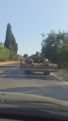 A vehicle is traveling on a paved road. The vehicle appears to be a tractor with a large agricultural attachment at the rear. A person is visible in the driver's seat of the tractor. The road is lined with trees on both sides, including tall cypress trees and olive trees. There is a speed limit sign indicating 50 on the right side of the road. The sky is clear and blue. The view is from another vehicle, looking forward.