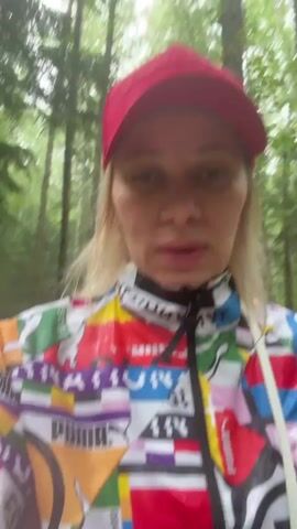 The video features a woman with blonde hair wearing a red baseball cap and a brightly colored Puma jacket. She is walking along a dirt path through a green forest. The woman is speaking, and appears to be upset that she is caught in the rain. The camera is held by the woman, creating a first-person perspective.