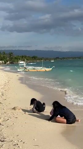 A video shows a sandy beach with a turquoise ocean. Two children, dressed in swimwear, are playing on the beach near the water's edge. The child on the left is partially obscured by sand and is in a crouching position. The child on the right is kneeling and also appears to be playing with the sand. In the background, several traditional outrigger boats are anchored in the clear water. The coastline is lined with palm trees and some structures. The sky is partly cloudy with a mix of blue sky and grey clouds. The waves are gently lapping onto the shore. The overall atmosphere suggests a serene beach setting.
