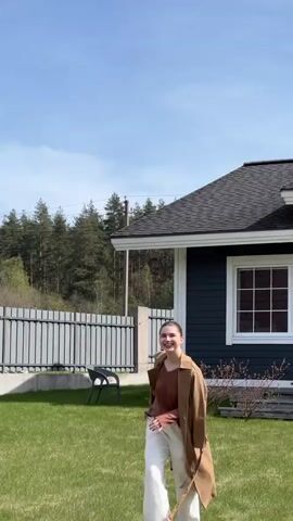 The video begins with a medium shot of a young woman with brown hair in a bun, wearing a brown long-sleeved shirt, white pants, and a tan trench coat, walking on a lawn toward the camera. She is smiling. A house is visible in the background to the right, with dark siding, white trim, and a gray roof. To the left of the house is a white fence and trees in the background. The sky is blue with a few clouds. The camera focuses on the woman as she comes closer and smiles, then the camera moves quickly away from her face and the focus is blurred.