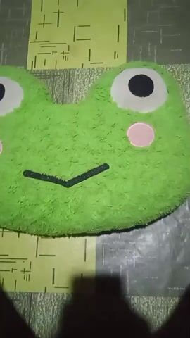 A close-up shot of a green, plush frog-shaped pillow. The pillow is lying on a patterned surface, possibly a table or floor, with yellow and gray lines. The frog has large white eyes with black pupils and pink circles on its cheeks. A black V-shaped mouth is also visible. The lighting is dim, and a shadow is cast in the foreground, obscuring the bottom part of the frame. The video appears to be a short, static recording with no discernible movement.