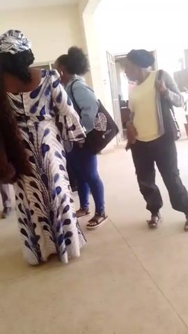 Three women are standing in a tiled room, possibly an indoor corridor, engaged in conversation. One woman is wearing a long white dress patterned with blue feather designs, a matching headwrap, and a brown shawl. Another woman stands beside her, wearing jeans, a white t-shirt, and a denim jacket. The third woman wears dark pants, a yellow t-shirt, and a dark head covering. She has a bag slung over her shoulder and is gesticulating as she talks. The overall impression is of a casual meeting or discussion taking place indoors.