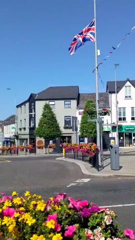 The video shows a street scene in a town with a Union Jack flag flying on a flagpole. The flag is positioned towards the right of the frame. In the foreground, there are colorful flowers, primarily pink and yellow. Behind the flowers, there is a roundabout with cars moving. There are several buildings lining the street, with shops on the ground floor and residential or office spaces above. One building is dark gray with a black roof, and another is white with a dark roof. There are hanging flower baskets on the buildings and along the street. A red car is seen turning. In the background, there is an archway decorated with circular images and flags. The sky is clear and blue, indicating a sunny day.