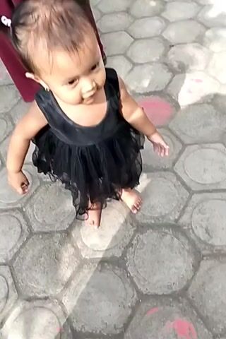 The video shows a young child, likely a toddler, standing on a patterned walkway made of hexagonal stones. The child is wearing a black, sleeveless dress with a tiered, sheer skirt and has their hair pulled up in two pigtails. They are looking to their right, with their hands clasped in front of their chest. The child's feet are bare. To the left of the frame, a person wearing a dark red garment is partially visible, and the lower legs of other individuals are visible in the background, suggesting a group is present. The walkway is illuminated by sunlight, creating shadows on the ground.