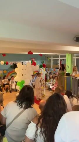The video depicts a graduation ceremony for young children, likely from preschool or kindergarten.  

A young boy in a cream-colored suit walks along a red carpet, approaching a woman in a tan jumpsuit who is handing him a small bouquet of green herbs or foliage. The boy has blonde hair and is smiling slightly. A decorated cardboard panel with cartoon figures stands behind him. 

A group of children sits on the floor to the left of the boy, some wearing small graduation gowns. Parents and adults are seated in chairs behind the camera's viewpoint, observing the ceremony.

The room is decorated with balloons, paper flowers, and a banner, suggesting a celebratory atmosphere. Another woman wearing a white blouse and grey pants stands near the first woman, holding an item that appears to be a small loaf of bread. The room is well-lit.