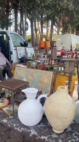 The video shows an outdoor market, likely a flea market or car boot sale. The first shot focuses on a table filled with various antique items, including pottery, vases, framed pictures, and decorative objects. The scene then pans to show people browsing through stalls and vendors setting up their wares. In the third shot, a man is holding a piece of what appears to be bark or wood, looking directly at the camera with a surprised expression. The final shot captures a wider view of the market, with stalls set up alongside parked vehicles. Several people are visible, some examining items, while others are interacting with vendors. The environment appears to be a paved area, possibly a parking lot or street, lined with trees. The weather seems to be sunny and pleasant.