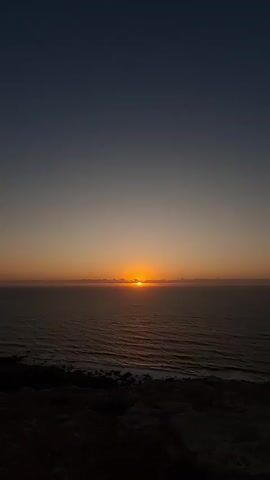 A serene sunset unfolds over the vast ocean, viewed from a rugged cliffside. The sun, a radiant orb of orange, hovers just above the horizon, casting a warm glow that bleeds into the sky and reflects upon the gently rippling water. Wispy clouds, tinged with pink and gold, stretch across the horizon, adding depth to the atmospheric scene. The ocean itself appears calm, with subtle waves lapping against the rocky shore below.

The foreground is dominated by dark, textured earth and stones, hinting at the elevated vantage point. The sky transitions from a deep, dusky blue at the top to softer hues of peach and orange as it nears the horizon, signifying the close of day. A few birds, mere specks against the expansive sky, add a touch of life to the tranquil panorama. The overall atmosphere is one of profound peace and natural beauty. The time of day is clearly evening, given the setting sun, and the weather appears clear, allowing for an unobstructed view of the celestial display. The location cues suggest a coastal area with dramatic cliffs. There is no visible text in the media file.