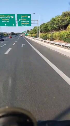 This media file captures a first-person perspective of driving on a highway. The primary subjects are the road, highway signs, passing vehicles, and the surrounding natural scenery. The setting is a multi-lane highway under a clear blue sky, suggesting daytime. The weather appears to be sunny and pleasant.

The activity taking place is driving, likely on a trip or commute. The camera is positioned as if mounted on a vehicle, possibly a motorcycle or scooter, given the close-up view of a dark, circular object in the foreground.

Notable details include several green highway signs. One sign, in Greek and English, indicates "Airport Services" and "Retail Park," along with "Cargo." Another sign in Greek and English lists "Flights-Arrivals," "Cargo," and "Military Base." The highway has clear white lane markings, and the road surface appears to be asphalt. Trees and vegetation line the right side of the highway, with a guardrail separating the roadway from the embankment. Several cars are visible on the highway, some in the distance and others closer to the camera.

The overall mood conveyed is one of travel and movement. The clear visibility and sunny conditions suggest a positive and uneventful journey. The location context identifies the area as the Paiania Municipal Unit in Greece, which aligns with the Greek text on the signs.