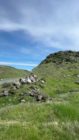 The video shows a sunny day in a grassy, hilly landscape. A road runs through the valley between the hills. There are power lines visible in the distance along the road. A pile of rocks sits in the foreground, with green grass covering the ground. The sky is blue with some scattered clouds and the sun is visible.