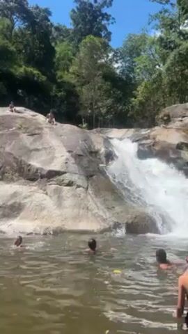 The video shows a small waterfall emptying into a pool of water where people are swimming. Several people are on the rocks at the top of the waterfall. One person slides down the rocks into the waterfall and drops into the pool. The setting is a natural area, with dense trees surrounding the waterfall and pool. The weather is sunny and the sky is clear.