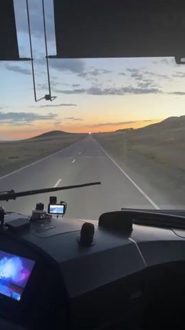 This is a first-person view from the driver's seat of a vehicle, likely a bus or truck, traveling down a long, straight road. The scene is set in a vast, open landscape of rolling hills under a dramatic sky. The time of day is clearly sunrise or sunset, with the sun appearing as a bright, distant point on the horizon. The sky is a gradient of blues, oranges, and yellows, with scattered clouds adding texture. The road is paved and appears to be in good condition, with clear lane markings.

In the foreground, within the vehicle's cabin, several objects are visible. On the dashboard, there is a screen displaying what appears to be a music video or concert footage, showing a woman performing on stage with bright lights and a cheering audience. Next to the screen are various electronic devices, including a small camera and a GPS unit. A microphone is also visible. The interior of the vehicle is dark, contrasting with the bright exterior scene.

The overall mood is one of travel and perhaps anticipation, given the expansive view and the colorful sky. The rural setting of Kazakhstan, specifically Baistau, suggests a journey through a natural and potentially remote area. The visible text on the screen in the cabin is not entirely legible but appears to be related to the performance shown. The motion of the vehicle is implied by the perspective and the clear road ahead.