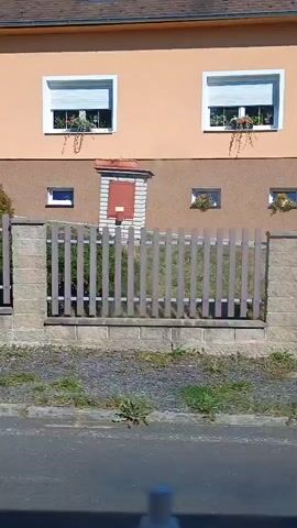The video shows a view from a moving vehicle, capturing residential properties and surrounding vegetation. The first frame shows a light orange house with a white window and a smaller brown window. A wooden picket fence with stone pillars separates the property from the street. The second frame shows a dark blue car parked on a driveway, with a metal gate. There are trees and bushes in the background, with a glimpse of a green fence. The third frame shows a white house with a dark roof, bordered by a wooden fence and green bushes. The fourth frame shows a white structure with stacked compartments, likely storage tanks, adjacent to a wooden fence and greenery. The final frame shows a yellow house with two windows adorned with flowers. A wooden fence and a stone wall are visible in front of the house. The lighting suggests it is daytime.