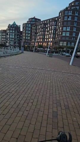 The video shows a person riding a bicycle along a paved walkway. The walkway is made of reddish-brown bricks and is lined with lampposts. In the background, there are modern brick buildings with many windows. To the left of the walkway, there is a harbor with several boats docked. The sky is cloudy and appears to be either sunrise or sunset. People are visible walking and cycling in the background.