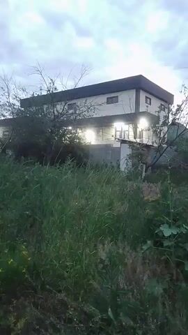 A two-story building with a dark roof and white walls is visible. There are illuminated windows on both floors. The building is partially obscured by trees and tall grass. The sky is cloudy. The video appears to be shaky, indicating movement.