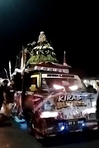 The video shows a decorated truck at night. The truck has a large, green, cone-shaped decoration on top, possibly made of leaves or foliage, with a figure or object at its very peak. The truck's headlights are on, and the front of the truck is decorated with graffiti and text. Flags are also visible on and around the truck. Several people are standing near the truck, some of whom appear to be holding onto or interacting with the decorations. The environment is dark, suggesting it is nighttime. The truck appears to be part of a procession or parade.