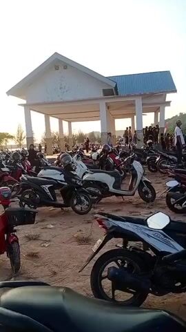 The video opens on a dirt parking lot filled with numerous motorcycles and scooters. In the background, a covered pavilion with white pillars stands against a pale sky. The sun is setting, casting a warm glow. To the left of the pavilion, trees and water are visible, suggesting a coastal or lakeside location. People are seen scattered amongst the vehicles and near the pavilion. The camera pans slowly to the right, revealing more trees, a road with some cars, and a few small tents or shelters near the water's edge, where more people are gathered. The overall scene depicts a public gathering or event at dusk in an outdoor setting, likely a recreational area.