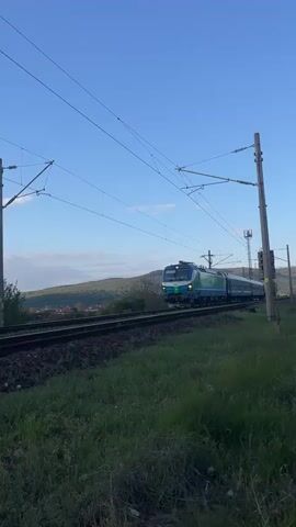 The video shows a blue and green train passing by on a track. The train, labeled "БДЖ BDZ" on the side, consists of a locomotive and several passenger cars, all moving from left to right in the frame. The locomotive has a number "902" displayed. Above the train, there are electrical lines supported by poles on either side of the track. The foreground consists of green grass, and in the background, there is a hill and a clear blue sky. The video is shot from a static position, at ground level.