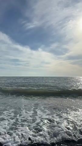 The video shows an ocean scene with small waves rolling in and out. The water is gray and foamy at the shoreline, becoming darker further out. The sky is mostly blue, filled with scattered clouds and the sun shining brightly. The overall impression is one of a calm, slightly overcast day at the beach.
