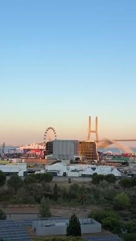 The video captures a wide shot of a festival or event setup with a large Ferris wheel and a prominent cable-stayed bridge in the background. The sky is a clear, pale blue, transitioning to a warmer hue near the horizon. Below the sky, the bridge's two distinctive H-shaped pylons and its network of cables are visible, spanning across a body of water. In the mid-ground, the event infrastructure includes several large tents, temporary structures, and what appears to be a stage or a large tent with a distinctive roof. There are numerous vehicles, including buses, vans, and cars, parked within the event grounds. A Ferris wheel, illuminated with lights, stands to the left of the bridge. The foreground features a natural landscape with trees and shrubs, and some solar panels are visible at the very bottom of the frame. The overall impression is of a large-scale outdoor event taking place during daylight or early evening, with the bridge serving as a significant landmark. There are no discernible individuals in the video.