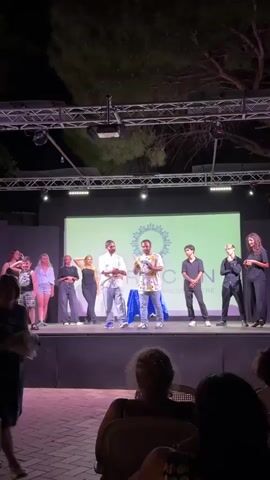 A group of people are on a stage in front of a large screen. The screen displays a logo with the word "IGNITION" and some text underneath. Two men in the center of the stage are interacting. One man, wearing a white shirt and blue jeans, is holding a microphone. The other man, wearing a colorful patterned shirt and blue jeans, is holding a blue object, possibly a toy or a prop, and appears to be demonstrating something. There are several other individuals on the stage, some standing and some in poses. In the foreground, out of focus, are several people seated, presumably an audience. The setting appears to be outdoors at night, with stage lighting illuminating the scene.