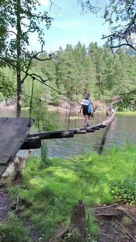 The video begins with a shot of a group of people crossing a narrow suspension bridge over a body of water. The bridge is made of wooden planks and rope. Lush green grass and trees surround the area. The camera then pans to a man with a beard, wearing a hat and backpack, who appears to be taking a selfie. A woman with a hat is visible in the background. The scene appears to be a hiking trail in a natural environment.