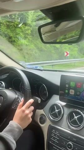 The video shows a woman driving a Mercedes-Benz, likely a CLA-Class, on a winding road. She is wearing glasses and a gray hoodie. The interior of the car is light tan and black. The rearview mirror is cracked. Music is playing. The driver appears to be activating or deactivating something on the driver's side door panel, perhaps a window lock or the child safety lock, while keeping her other hand on the wheel. Lush green trees line the road outside.