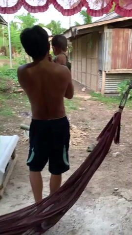 A shirtless person wearing shorts and flip-flops is holding a small child. The person is facing away from the camera and towards a building made of wood and corrugated metal. The building appears to be in a rural setting, with vegetation and dirt visible in the background. A hammock is hanging in the foreground.