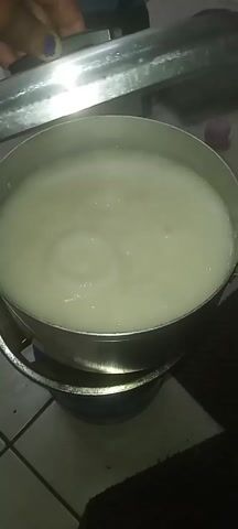 The video shows a large metal pot sitting on a gas stove. Inside the pot is a white, semi-liquid substance that is bubbling. A person's hand is visible holding a metal lid slightly ajar over the pot. The stovetop is made of white tiles. Steam is rising from the pot.