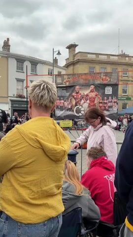 The video captures an outdoor event, possibly a street fair or competition, with a crowd of people gathered. In the foreground, a person wearing a bright yellow hooded jacket is seen from behind. To their right, another person in a pink hoodie is leaning over a railing, looking down. Behind them, a group of people are seated in chairs, also facing the event. In the background, a large banner displays images of muscular individuals, suggesting a bodybuilding or strength-related competition. A stage or performance area is visible, with scaffolding and other structures in the vicinity. The overall environment appears to be an urban setting with buildings lining the street. The atmosphere seems casual and observational, with people watching the unfolding event.
