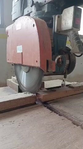 The video shows a close-up view of a large radial arm saw cutting a piece of wood. The saw blade is spinning at high speed, and sawdust is visible. The wood is held in place by clamps on a wooden workbench. The radial arm saw is positioned above the wood, and the blade is descending to make the cut. The overall environment appears to be a workshop or construction site, with the machine and raw materials being the primary focus.
