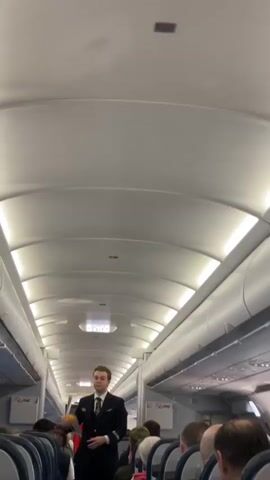 The interior of an airplane is shown with an attendant standing in the center aisle facing the back of the plane giving instructions while holding his arms open.  He is wearing a black suit and light-colored tie. Passengers are seated in their seats, and overhead bins line the top sides of the plane.