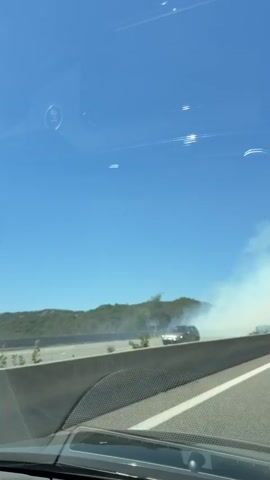 The video captures a view from inside a vehicle traveling on a highway. A concrete barrier separates the highway lanes from the roadside vegetation. On the right side of the highway, a plume of white smoke is visible rising from the vegetation, indicating a potential fire. In the distance, rolling hills covered in trees and vegetation can be seen under a clear blue sky. Two cars are visible on the highway, one in the foreground on the left and another further ahead on the right, both moving in the same direction as the camera. The smoke appears to be spreading across the roadside.