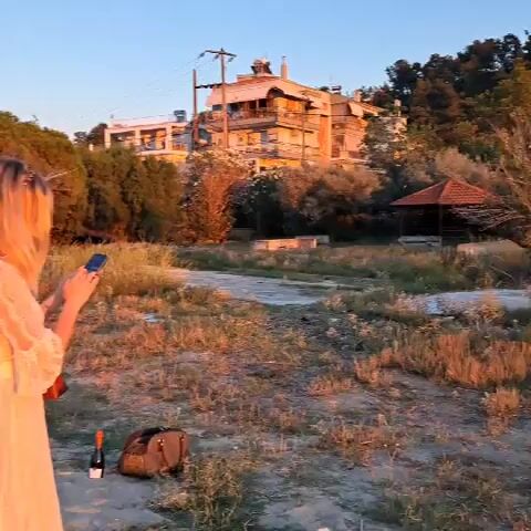 The video shows a group of women on a beach at sunset. The woman in the foreground, facing away from the camera, is wearing a light-colored dress and holding a phone, possibly taking a picture. In the background, several other women are standing near the water, some wearing white dresses. There are picnic items on the sand, including a blanket, bags, and bottles. The setting appears to be a coastal area with trees and buildings visible in the distance. The sky is a gradient of blue and pink, indicative of sunset.