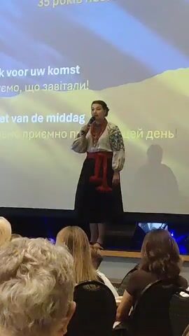The video shows a woman singing into a microphone on a stage. She is wearing traditional Ukrainian clothing, including an embroidered shirt and a black skirt with a red sash. Behind her, a projection screen displays text and a yellow and blue image, resembling the Ukrainian flag. In the foreground, the audience is visible, with some individuals' heads and shoulders in view. The lighting is dim, with the spotlight focused on the singer. The overall atmosphere appears to be a cultural event or performance.