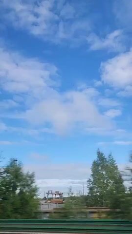 The video captures a scene of a drive-by perspective on a partly cloudy day. The camera is positioned inside a moving vehicle, offering a view of the sky and the landscape through the window. The sky is a bright blue, dotted with patches of fluffy white clouds. As the vehicle moves, trees in varying shades of green blur past, suggesting a relatively high speed. Structures like utility poles, a tall metal tower, and a long, light-colored building with multiple windows and a chimney are visible in the background. A green guardrail runs along the bottom of the frame, indicating a roadside view. The overall impression is one of observing a semi-urban or industrial landscape while in transit on a clear day.