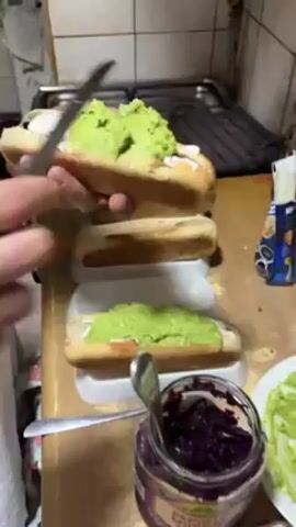 A person is making a hotdog. The individual is spreading guacamole on top of the hotdog with a serrated knife. The hotdog has a white sauce and red cabbage already on it. Other hotdogs with the same toppings can be seen on the cutting board nearby. There is a jar of red cabbage and a container of guacamole next to the cutting board. The scene is a kitchen with a stove and oven in the background.