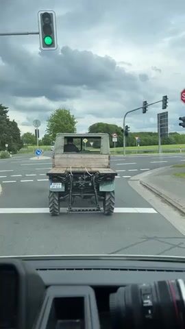 The video shows the back of a vintage, green farm tractor with a wooden platform and canopy, stopped at a traffic light. The driver is visible, sitting inside. The tractor has large, treaded tires, and a license plate that reads "SY UM 411". The background includes trees, a roundabout, and a cloudy sky. The video is shot from inside a car, with a camera lens visible in the foreground. The traffic light turns green, and the tractor begins to move.